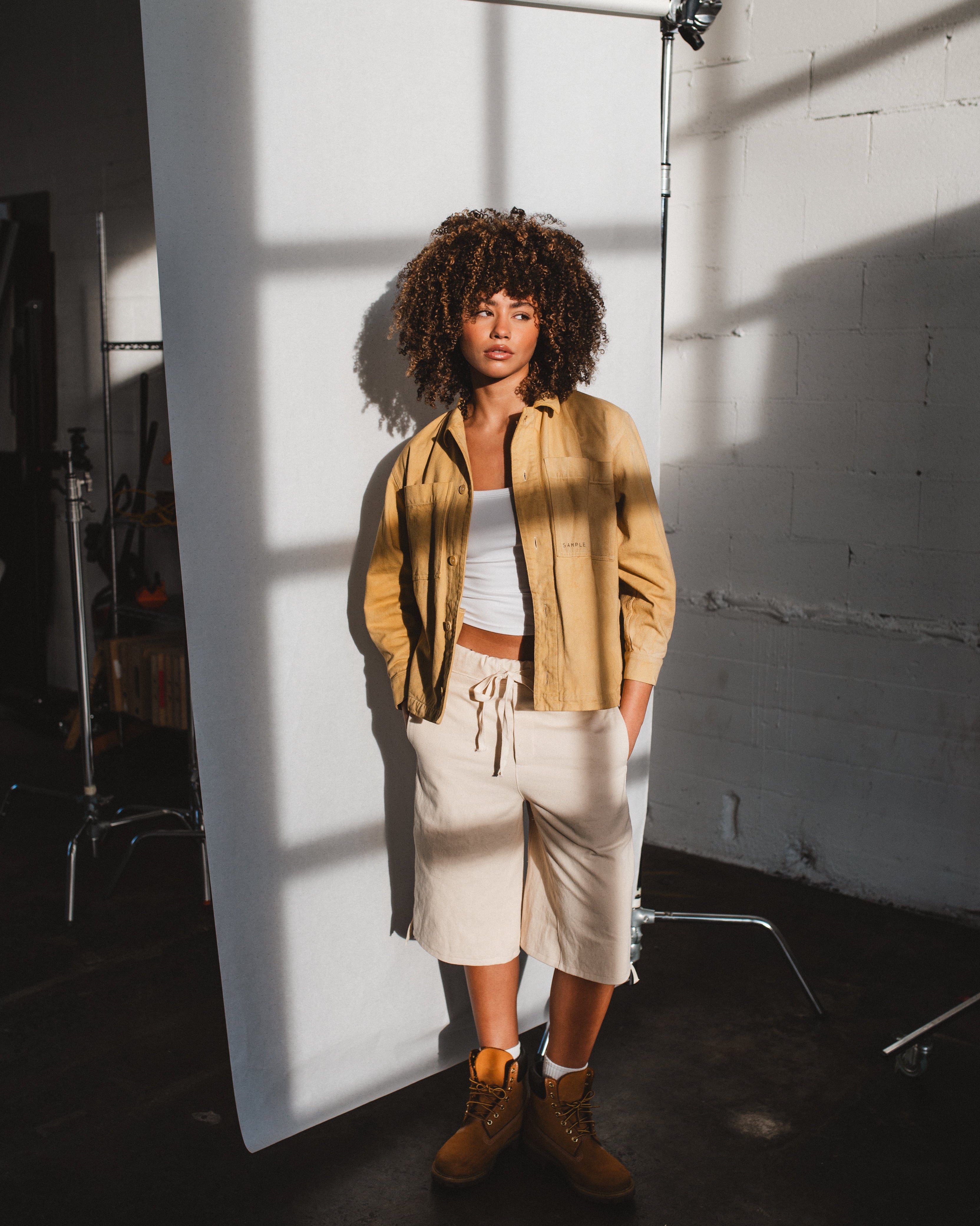 Person standing in a studio with a white backdrop, wearing a beige shirt, white pants, and brown shoes.