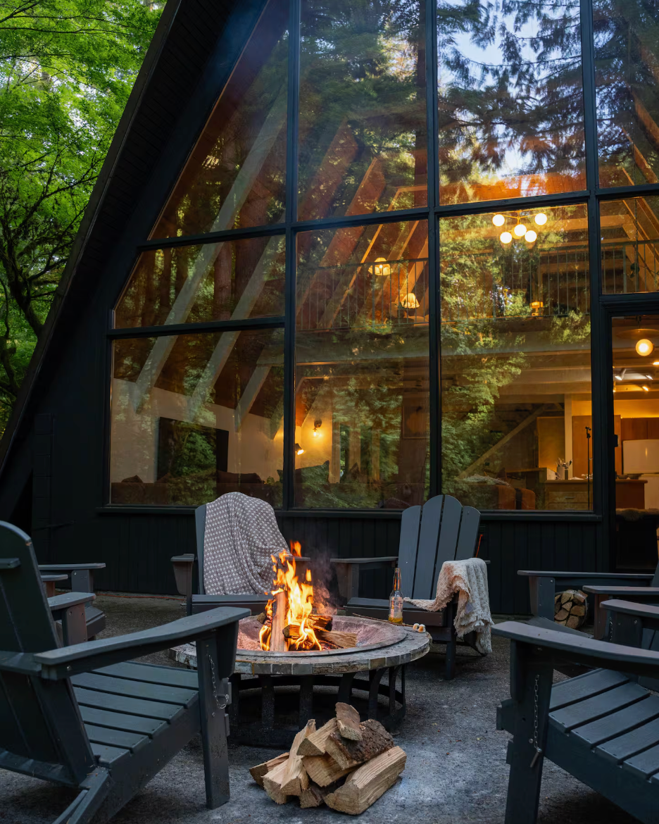 Outdoor setting with a fire pit and Adirondack chairs in front of a glass-walled building.