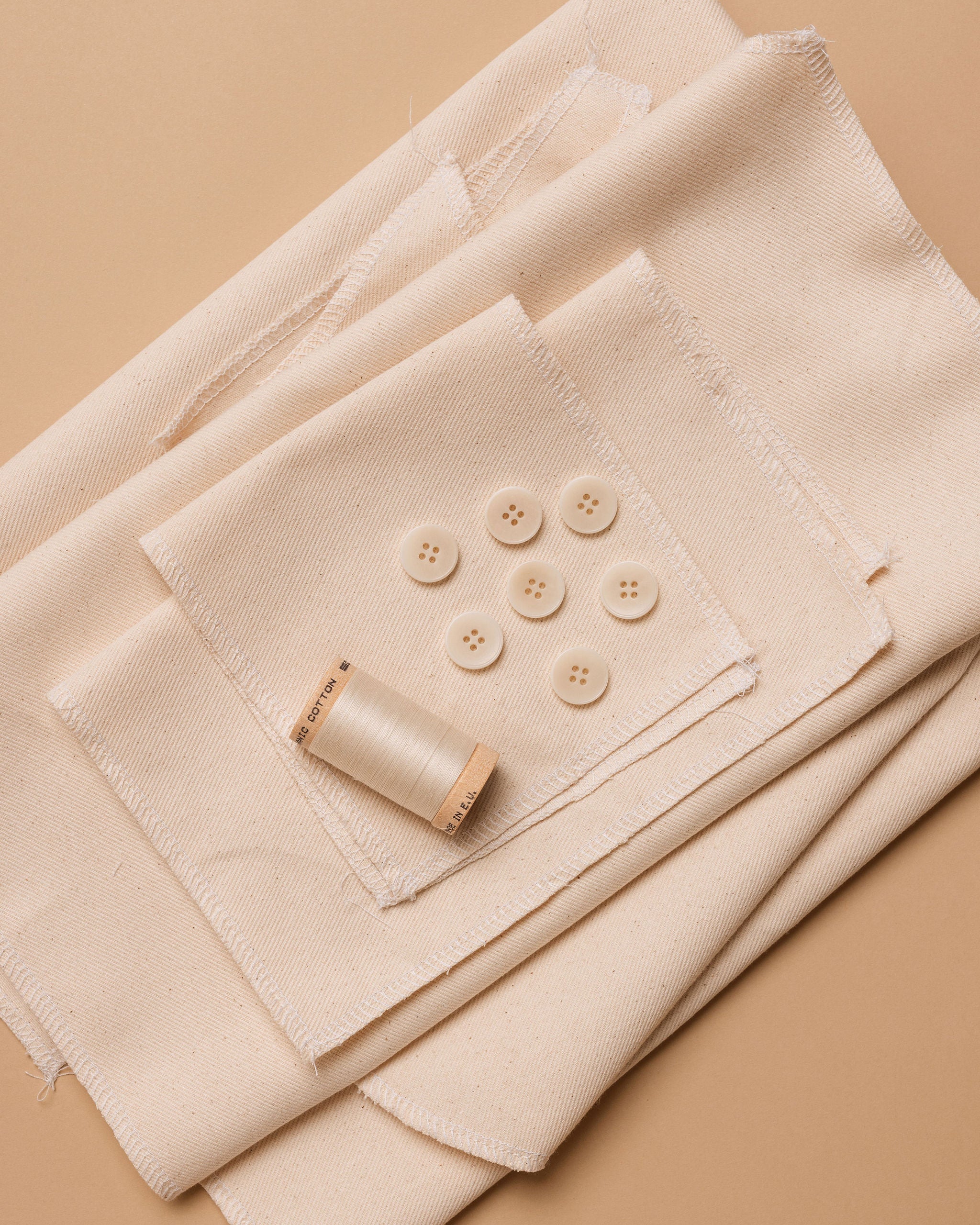 A collection of beige colored fabric pieces laid out flat, accompanied by a spool of thread and buttons.