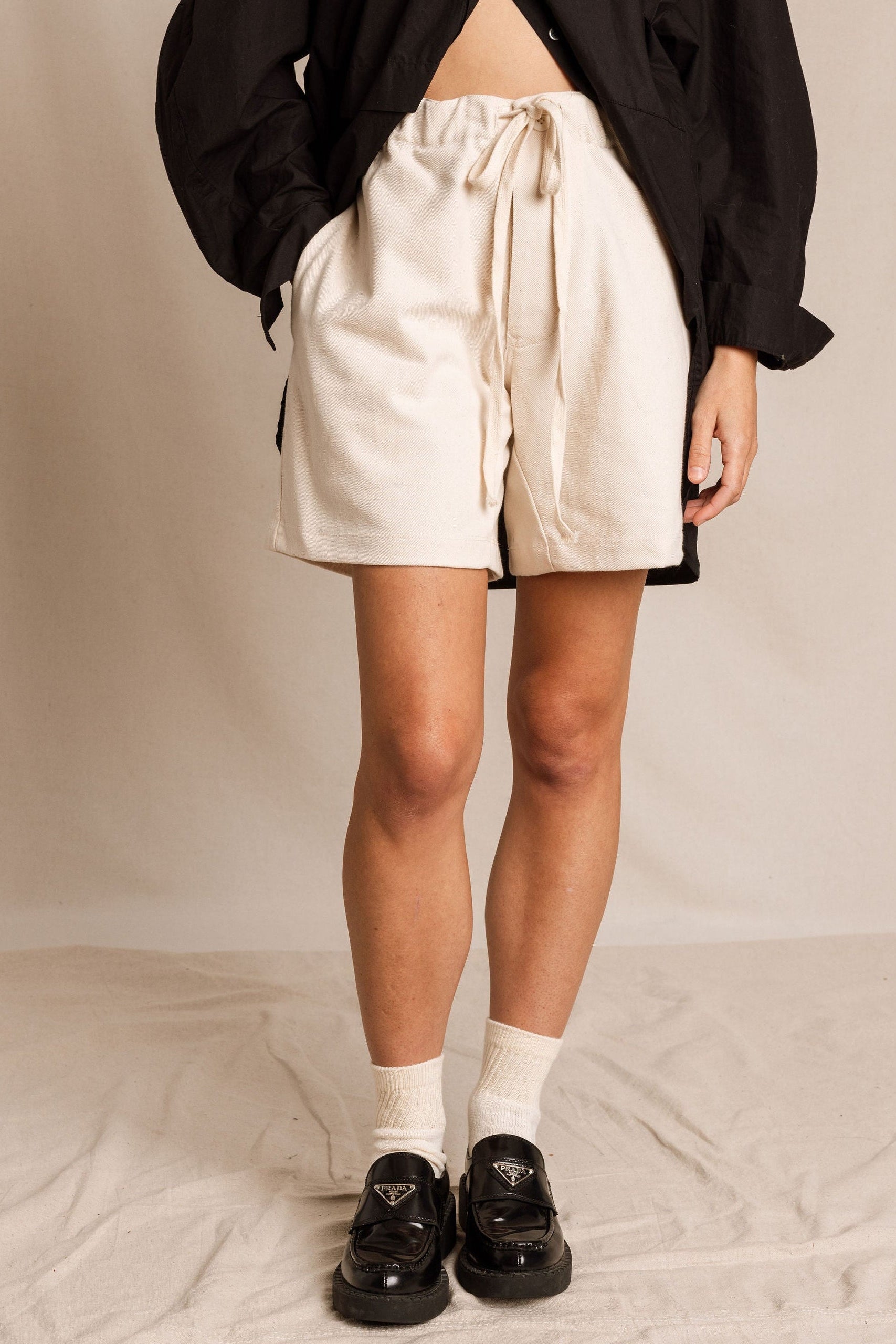 Person wearing beige shorts, black shirt, and black shoes on a neutral background